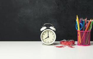 white round alarm clock and a metal glass with pens, pencils and felt-tip pens on the background of an empty black chalk board photo