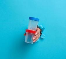 female hand holds an empty plastic jar for urine tests, the hand is sticking out of a torn hole in a blue background photo