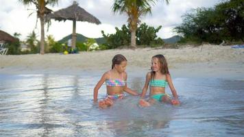 adorable pequeño muchachas jugando con arena en el playa. niños sentado en superficial agua y haciendo un arena castillo video