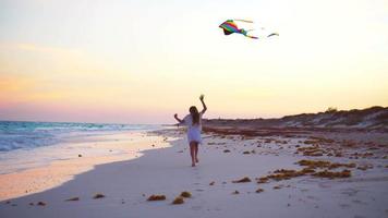 niña corriendo con cometa voladora en la playa tropical al atardecer. los niños juegan en la orilla del océano. niño con juguetes de playa. video