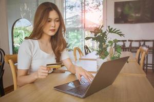 joven asiática con tarjeta de crédito y usando computadora portátil. hermosa joven feliz en el café. compras en línea, comercio electrónico, gasto de dinero, billetera de dinero tecnológico y concepto de pago en línea. foto