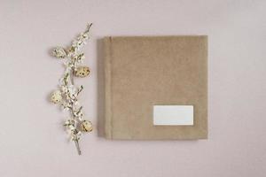 Flat lay with a beige photo album or book with a metal frame for the inscription, spring branches with white flowers and Easter eggs on a beige background. Top view, copy space