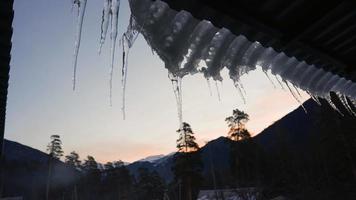 detailopname. bevroren ijspegels Aan de rand van de dak Aan de achtergrond van een berg landschap in de avond. winter tijd. langzaam beweging. video