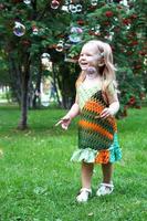 una linda niña sonriente con coloridos vestidos de verano está jugando con pompas de jabón en el parque de verano. foto