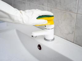 a hand in a white rubber glove with a sponge washes a top of a white faucet photo