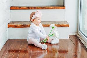 Little smiling baby girl one year old wearing spring wreath siting on floor in bright light living room near window and playing with gerbera flowers. Happy kid playing at home. Childhood concept. photo