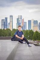 The boy is sitting on the steps in the open air against the background of skyscrapers and high-rise buildings. Journey. Lifestyle in the city. photo