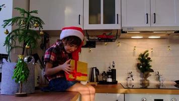 niña feliz está indignada, jura y amenaza con el puño que la caja está vacía y allí y muecas graciosas. niño con sombrero de santa está sentado en una cocina festiva decorada para navidad. Año Nuevo video