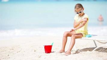 liten flicka med flaska av Sol grädde Sammanträde på tropisk strand video