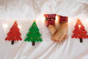 Kid bare feet Christmas garland lights background. Child in pajamas sleep on white cozy bed holiday night.Hand made paper garland red and green photo