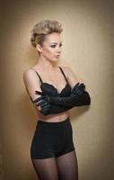 Attractive fair hair model with pantyhose and black bra and long gloves posing provocatively. Fashion portrait of sensual blonde, studio shot. Sensual female in black lingerie posing against wall. photo