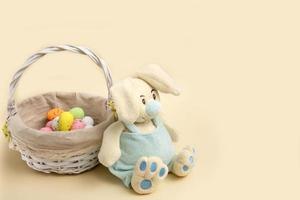 easter bunny sits by a basket with colorful eggs on a beige background with copy space photo