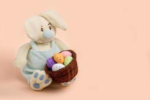 toy bunny with a basket of colorful Easter eggs on a peach background with copy space photo