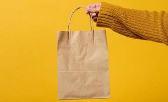 female hand in a sweater holds a brown craft paper bag with handles on a yellow background. Zero waste photo