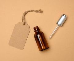 paper tag and glass cosmetic brown bottle with a pipette on a beige background. Cosmetics SPA branding mockup, top view photo