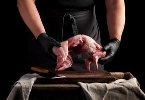 chef in black latex gloves holds a whole rabbit carcass over a brown cutting board, meat cooking process photo