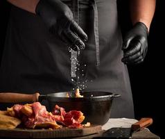 chef in black uniform and latex gloves sprinkles with white salt raw chicken meat in a black cast-iron frying pan, cooking photo