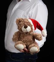 man in a white shirt holds a teddy brown bear in a red cap photo