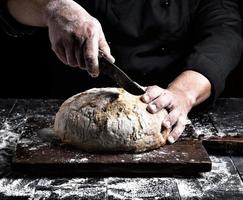 man cuts with a knife a round whole loaf of white wheat flour photo