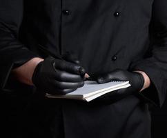 chef in black uniform and latex gloves holding a blank notebook and a black wooden pencil photo