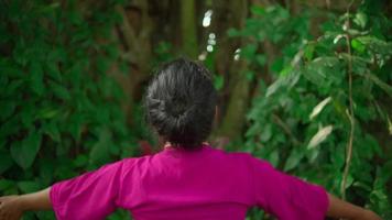 une belle femme asiatique fait un rituel de danse tout en portant une robe rose et un maquillage devant le grand arbre plein de buissons verts à l'intérieur du village video