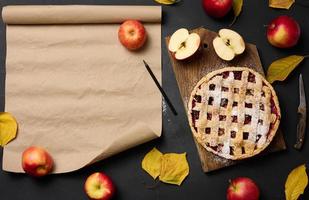 baked round traditional apple pie on a brown wooden board and fresh red apples. Nearby is a roll of brown paper for writing a recipe or menu photo