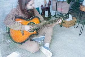 joven mujer jugando guitarra a hogar, ver mediante el vaso ventana foto