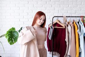 young beautiful woman wearing beige pastel coat, standing near clothes rack. Wardrobe change photo