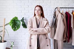 young beautiful woman wearing beige pastel coat, standing near clothes rack. Wardrobe change photo