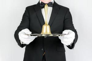 Portrait of Butler or Waiter in Dark Suit on White Background Holding Gold Bell on Silver Tray. Concept of Service Industry and Professional Hospitality. photo