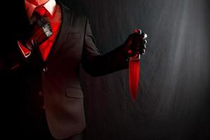 Portrait of Businessman in Dark Suit Holding Bloody Red Knife on Black Background. Concept of Horror Movie Killer and Stylish Assassin. photo