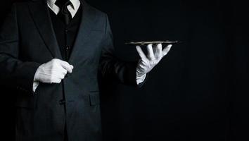 Portrait of Butler or Waiter in Dark Suit and White Gloves Holding Serving Tray on Black Background. Copy Space for Service Industry and Professional Hospitality. photo