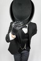 Man in Dark Suit and Leather Gloves Politely Doffing Bowler Hat on White Background. Concept of Classic and Eccentric British Gentleman Stereotype. Retro Style and Vintage Fashion. photo