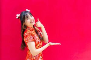 Cute Asian girl with long hair who wears a red Cheongsam dress in Chinese new year theme while she shows her hand to present something on a red background. photo