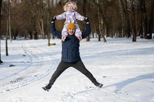 Father hold child on shoulders while jumping together at sunny frosty winter day in the park. Dad and daughter love. photo