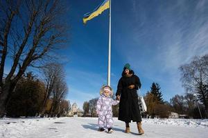 Mother and child walking on a sunny frosty winter day in the park, against background of a flagpole with the Ukrainian flag. photo