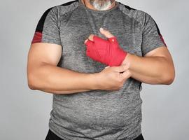 athlete is wearing gray clothes and wrapping his hands in red textile elastic bandage photo