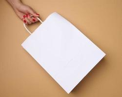 female hand holds an empty white paper bag by the handles on a brown background, rejection of plastic bags photo