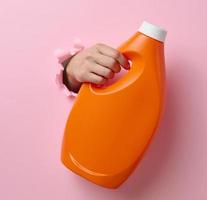 orange plastic bottle with liquid detergent in a female hand on a pink background. A part of the body sticks out of a torn hole in the background photo