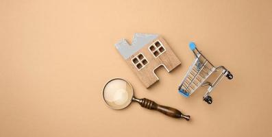 model of a wooden house, a miniature shopping cart and a magnifying glass on a light brown background. Home search concept for rent, purchase, mortgage photo