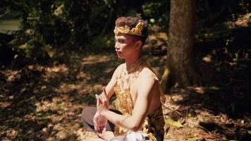 An Asian man with a golden crown sitting on a stone while meditating in the forest in a shirtless video