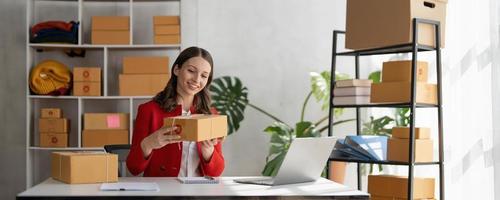 Happy young asian woman startup small business freelance holding parcel box and computer laptop and sitting on chair, Online marketing packing box delivery concept photo