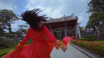 un' bellissimo giovane Cinese donna giocando con un' rosso cheongsam vestito dentro il giardino video