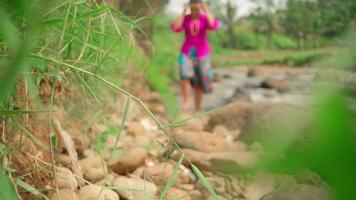 Asian woman walking on the edge of the river while wearing a pink dress and green skirt in makeup with a lot of rock video