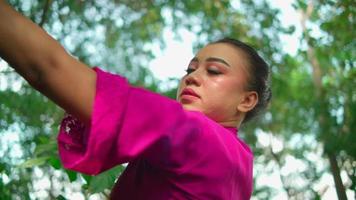 una hermosa mujer asiática hace un ritual de baile mientras usa un vestido rosa y maquillaje frente al gran árbol lleno de arbustos verdes dentro del pueblo video