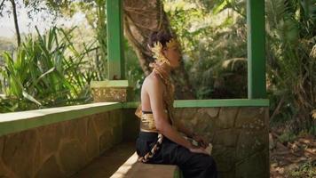 balinesischer mann, der friedlich sitzt, während er in einer goldenen krone und einem goldenen kostüm im dschungel meditiert video