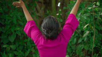 A beautiful Asian woman does a dancing ritual while wearing a pink dress and makeup in front of the big tree full of green bushes inside the village video