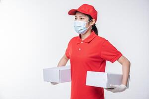 imagen de un joven repartidor consciente con gorra roja en blanco, camiseta uniforme, guantes de máscara facial de pie con una caja de cartón blanca vacía aislada en un estudio de fondo gris claro foto