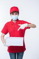 imagen de un joven repartidor consciente con gorra roja en blanco, camiseta uniforme, guantes de máscara facial de pie con una caja de cartón blanca vacía aislada en un estudio de fondo gris claro foto