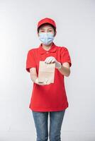 imagen de un joven repartidor feliz con gorra roja en blanco camiseta uniforme mascarilla guantes de pie con un paquete de papel artesanal marrón vacío aislado en un estudio de fondo gris claro foto